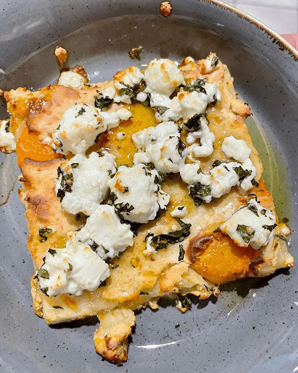 A dish with a square flatbread with feta cheese and herbs over it.