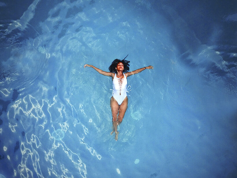 Aerial shot of a woman in white bathing suit floating with her eyes closed in blue waters.