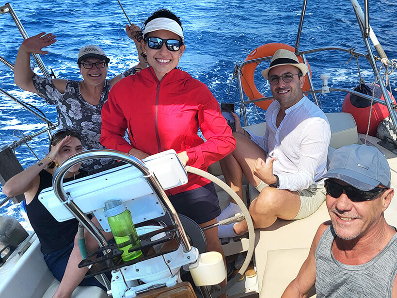 Stefania behind the helm of a sailing yacht with happy guests around her in the cockpit while sailing.